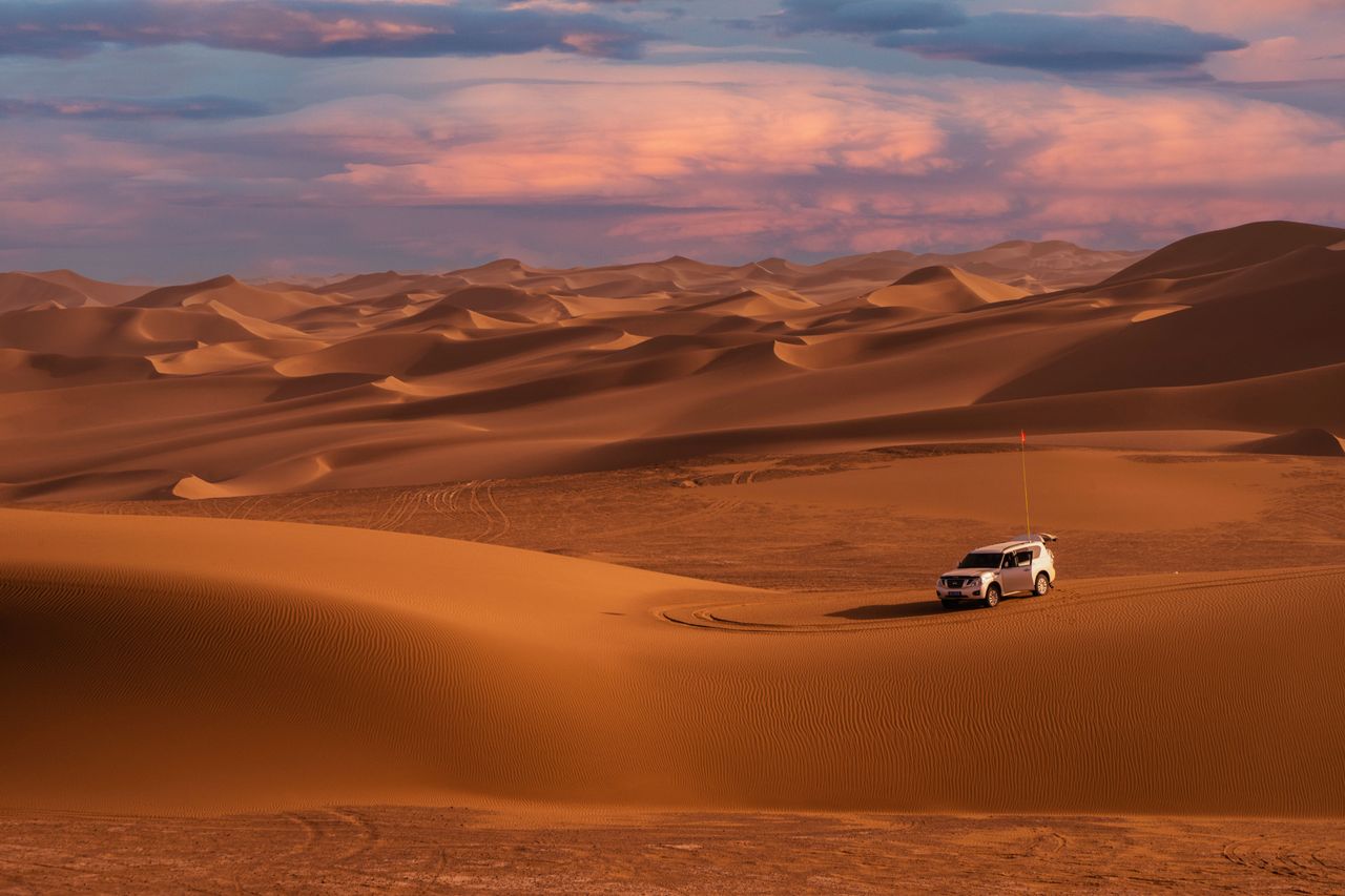 A vehicle traverses vast desert dunes under a beautiful sunset sky, showcasing the rugged landscape.