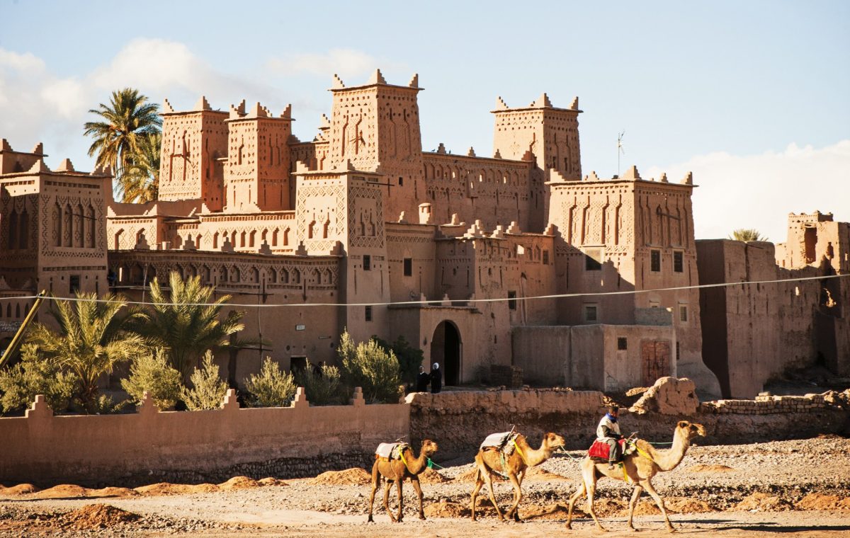 Earthen kasbah with palm trees and camels walking past, on the road from Fes to Marrakech